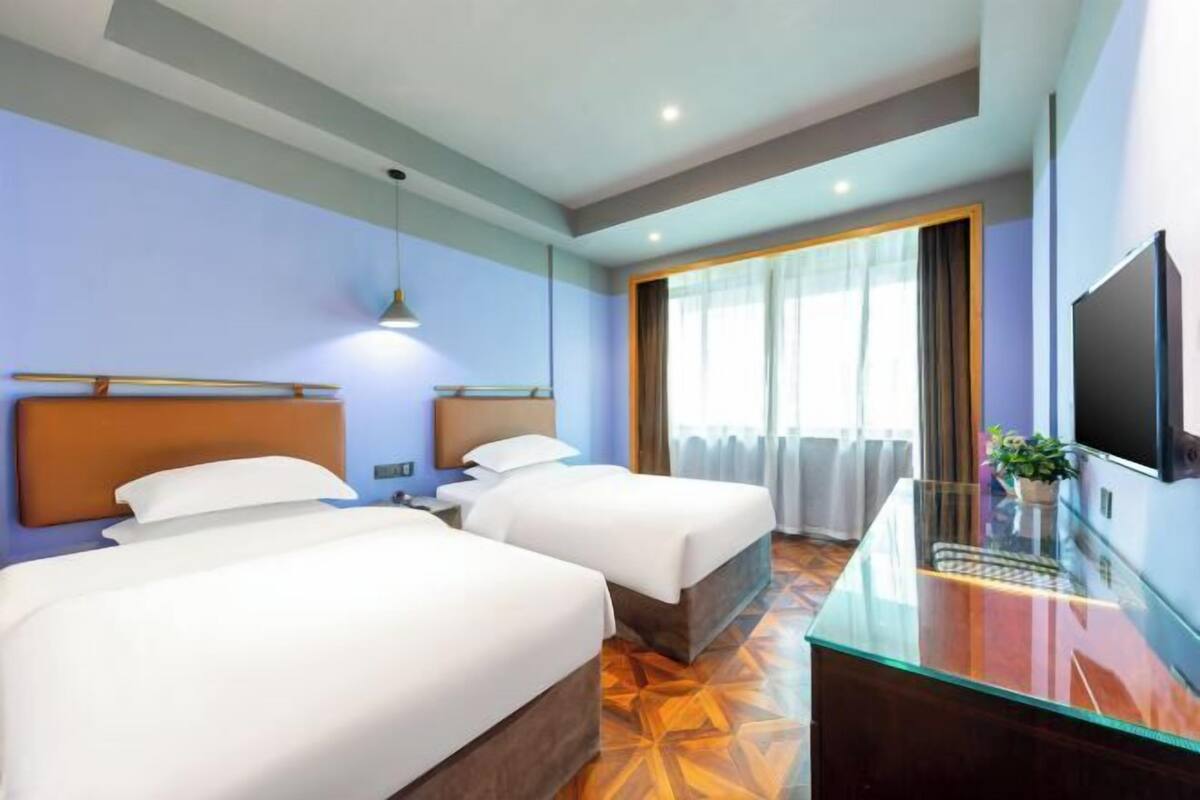 A hotel room with two beds, a glass desk, a TV, and a large window with curtains.