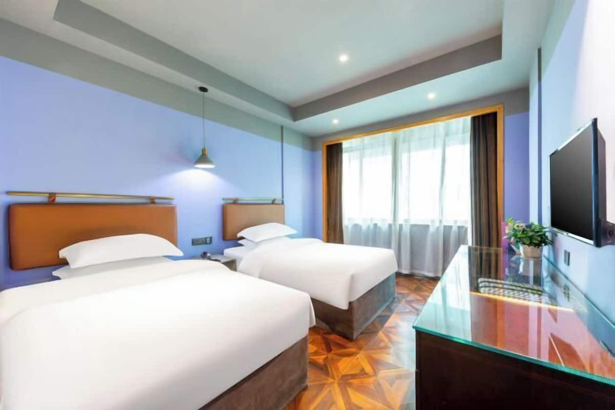 A hotel room with two beds, a glass desk, a TV, and a large window with curtains.
