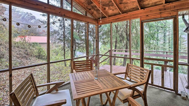 Une terrasse en bois avec une table et des chaises, de grandes fenêtres et une vue sur des arbres et un bâtiment au toit rouge.