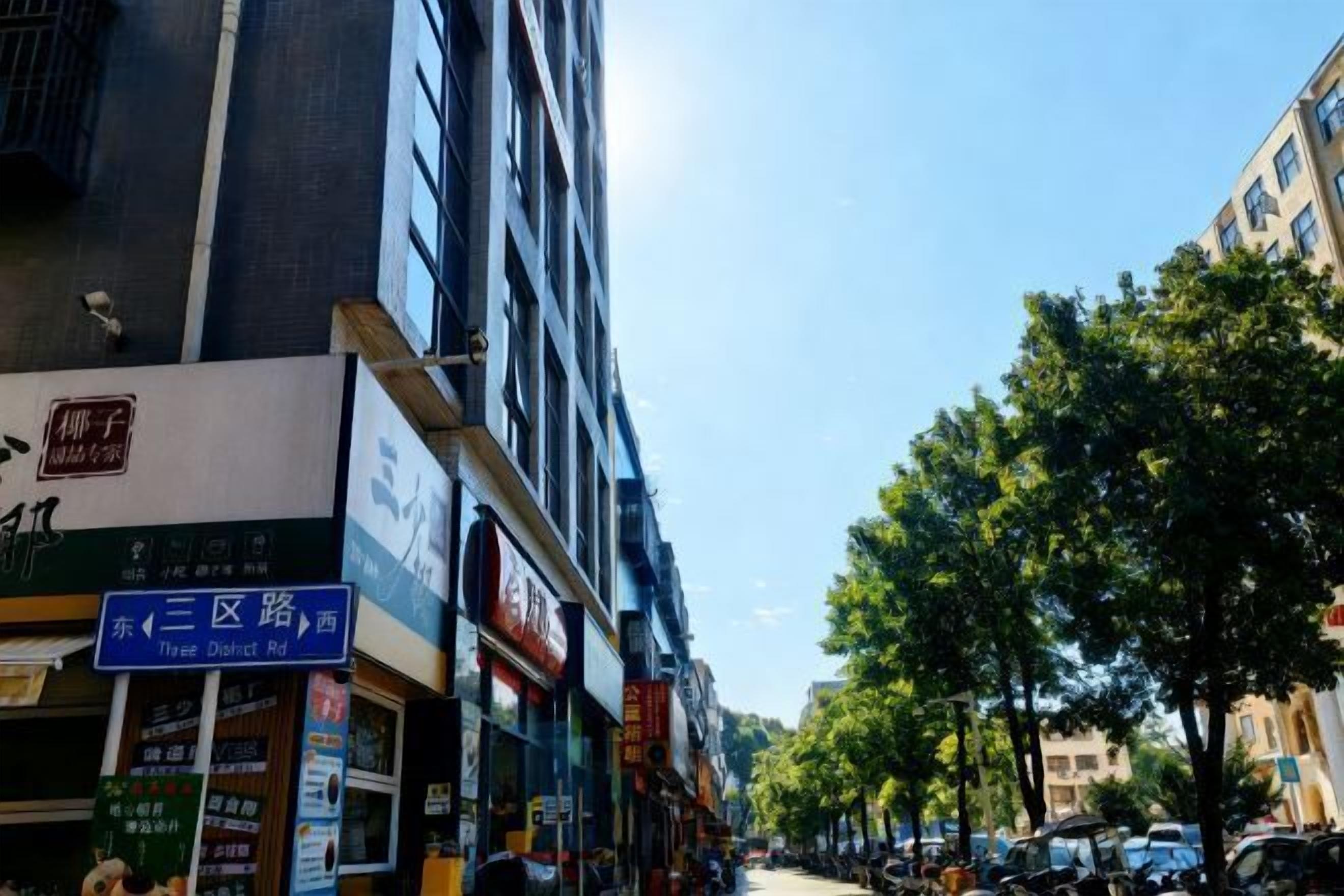 Une vue de rue avec un immeuble de trois étages, une enseigne indiquant « Three District Rd », et des voitures garées le long du trottoir.