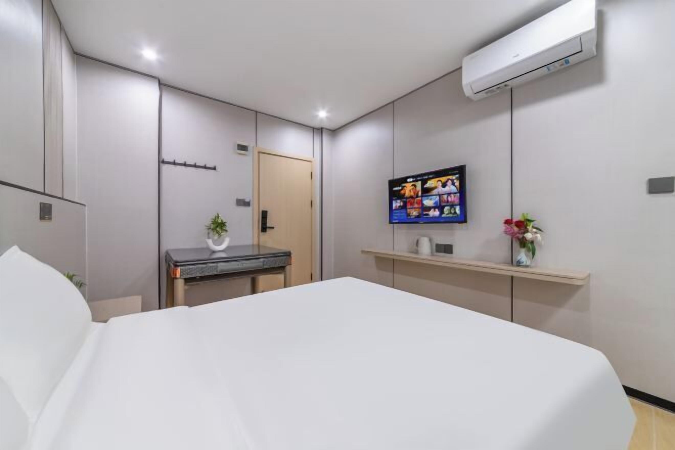A modern hotel room with a large bed, a flat-screen TV mounted on the wall, a small desk with a plant, and a shelf with a vase of flowers.