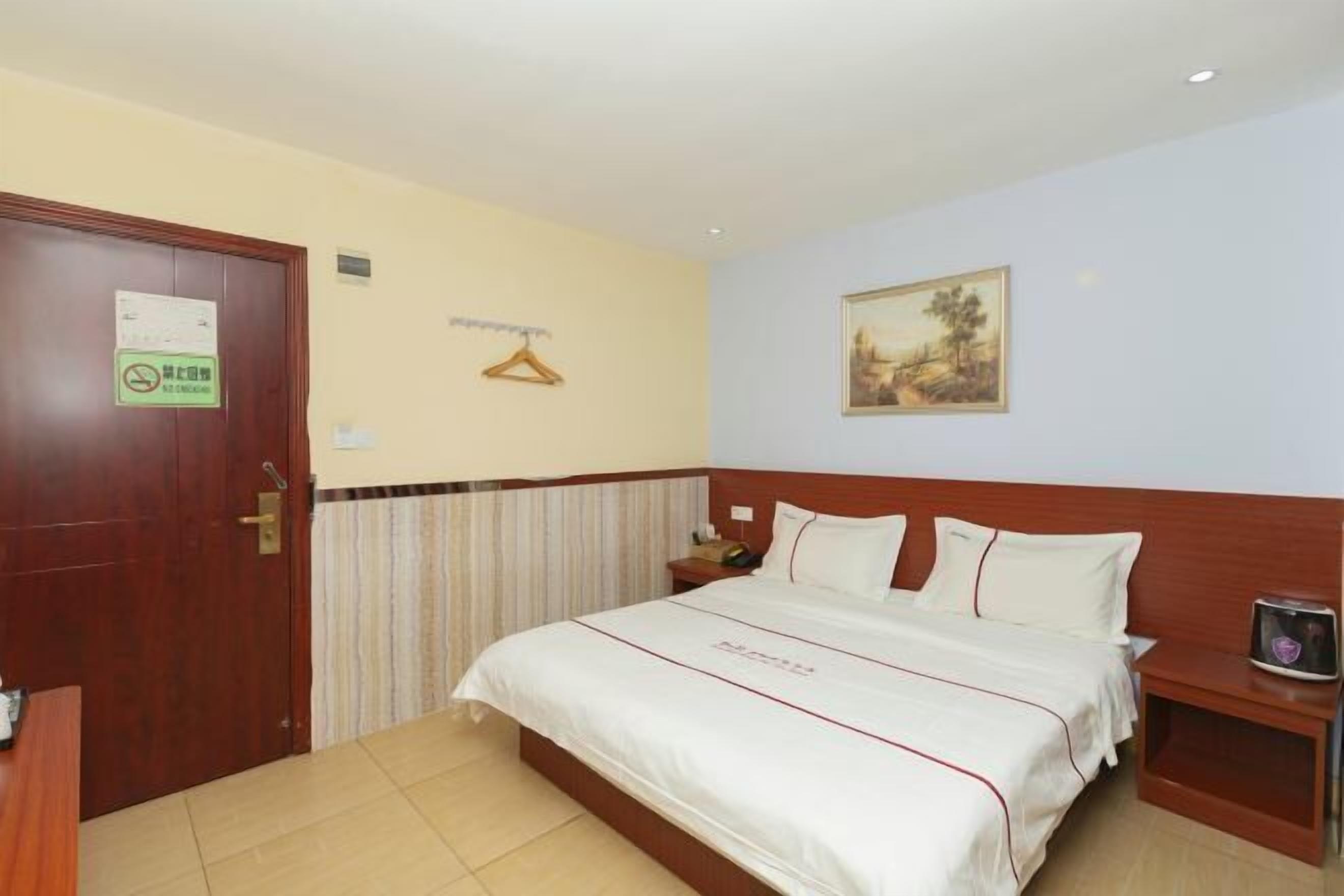 Habitación de hotel con una cama, una puerta de madera, perchero, un cuadro en la pared y una pequeña mesa con un teléfono y un adorno.