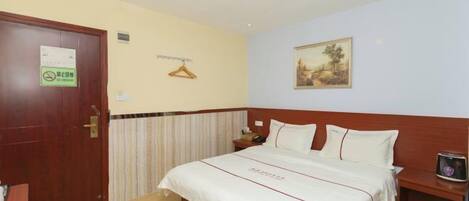 Habitación de hotel con una cama, una puerta de madera, perchero, un cuadro en la pared y una pequeña mesa con un teléfono y un adorno.