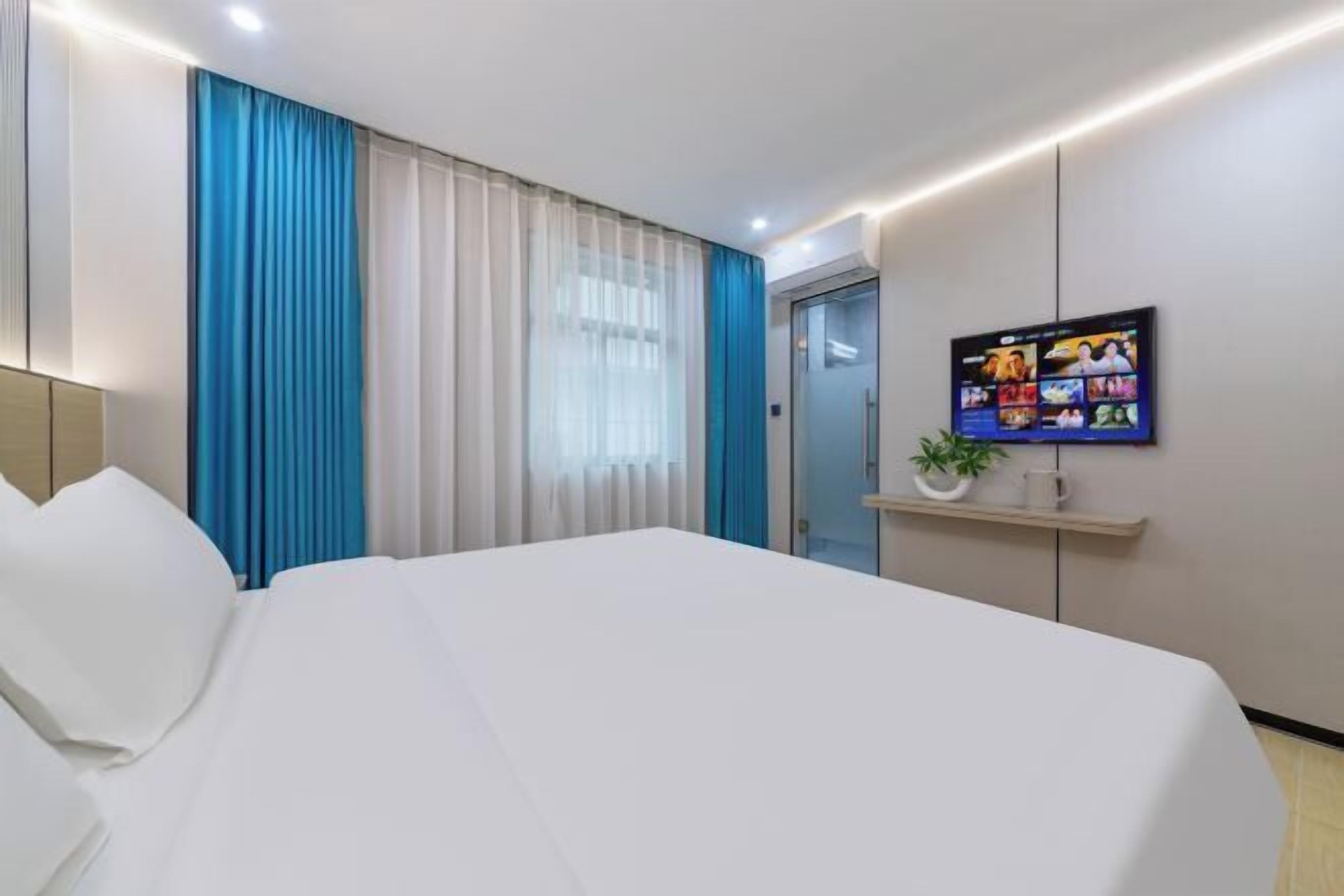 A hotel room with a large bed, a television mounted on the wall, and a small shelf with a plant and a mug.