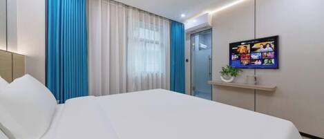 A hotel room with a large bed, a television mounted on the wall, and a small shelf with a plant and a mug.