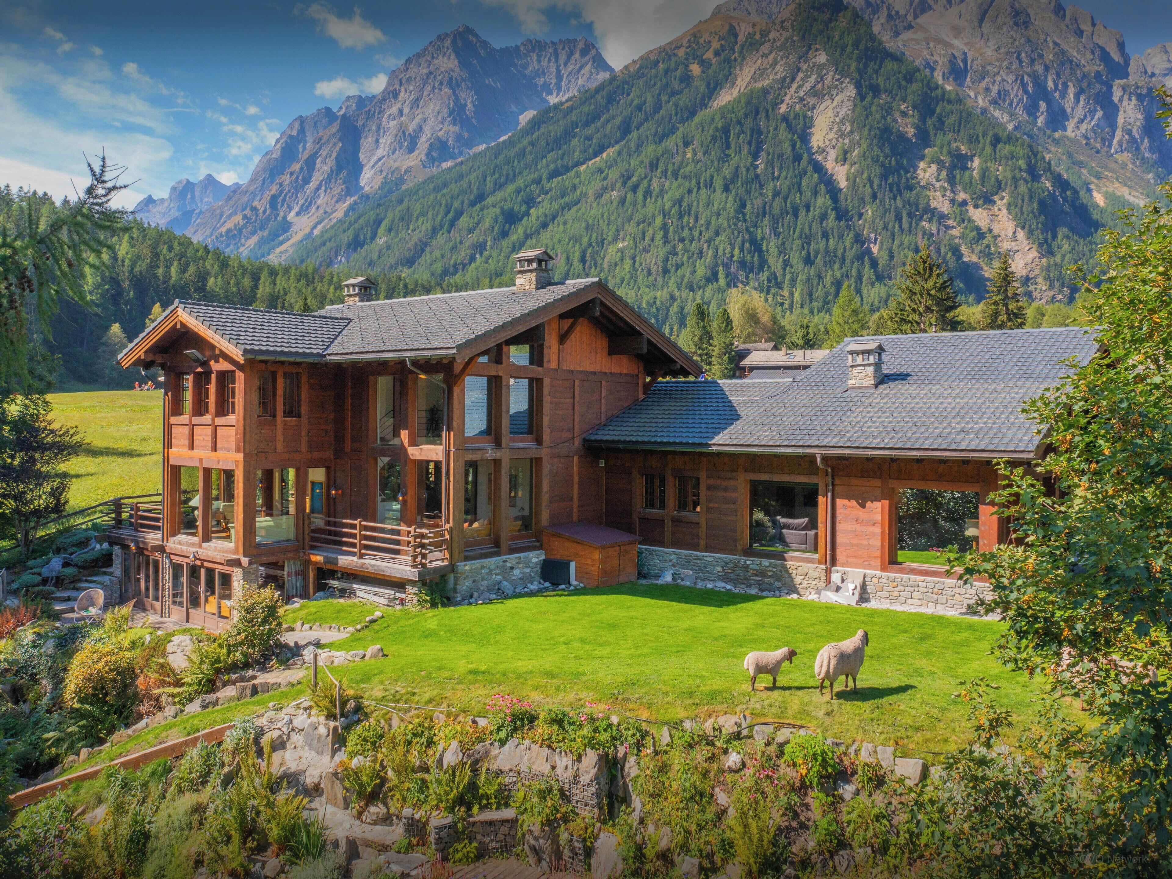 A wooden chalet with a stone foundation, large windows, and a balcony overlooking a grassy area with sheep.