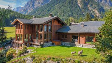 A wooden chalet with a stone foundation, large windows, and a balcony overlooking a grassy area with sheep.