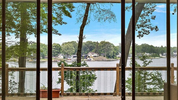 A room with a large sliding glass door leading to a deck with a view of a lake and trees. There are two sofas and a small table.