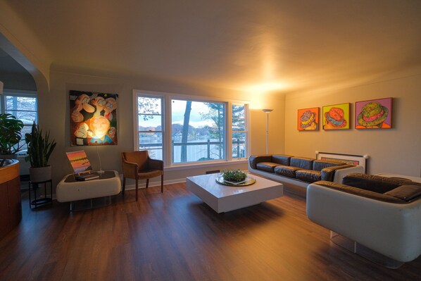 A modern living room with a white sofa, a coffee table, and a large window with a view. There are three paintings on the wall and a potted plant. - Family Compound: waterfront, 6 acres + pool (Spring Lake)