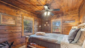 A wooden cabin bedroom with a bed, a ceiling fan, and a 'STAY awhile' sign on the wall.