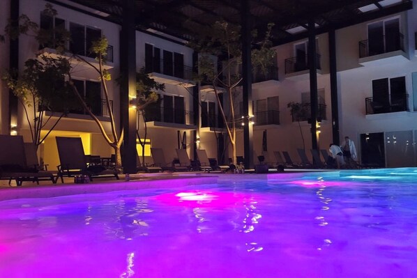 A swimming pool with colorful lighting, surrounded by lounge chairs and trees, with a building in the background.