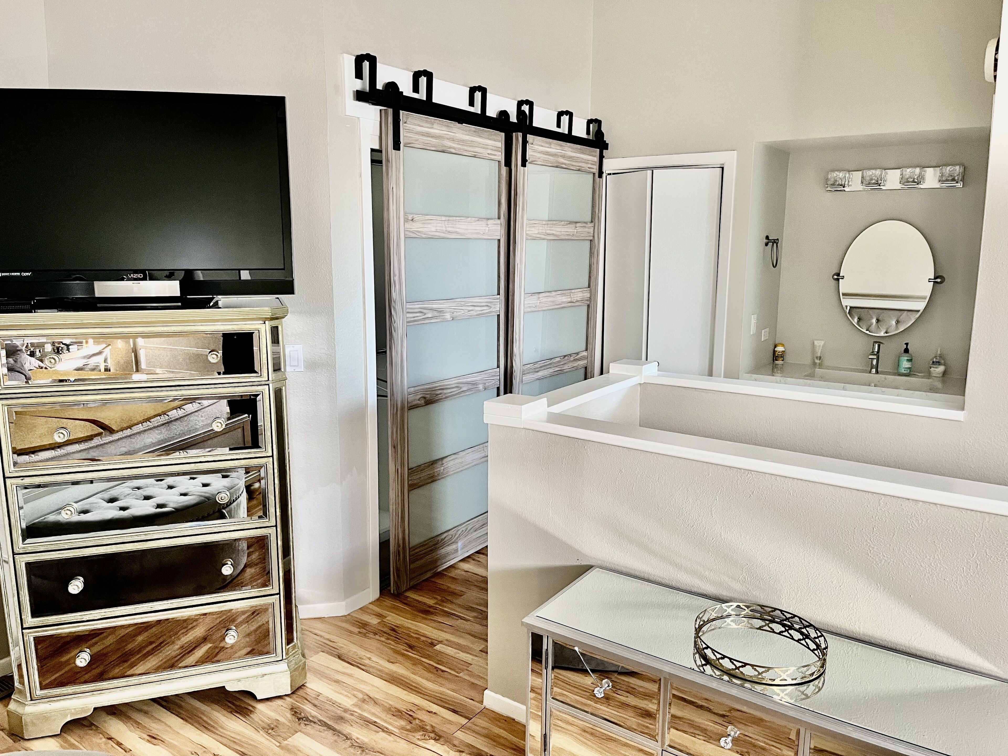 A room with a television on a stand, a sliding door, a bathroom with a mirror and sink, and a small table with a decorative item.