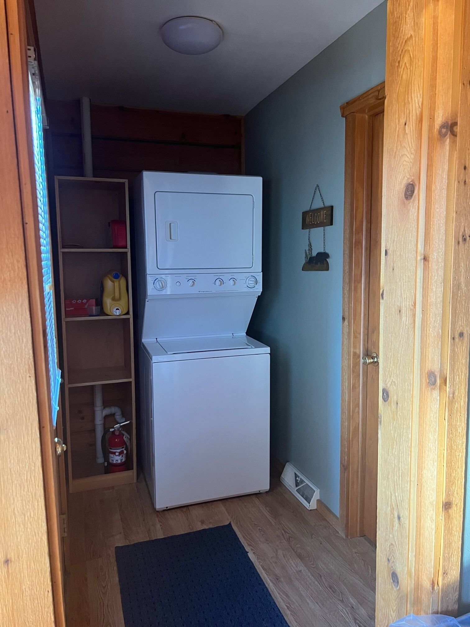 A laundry room with a stacked washer and dryer, wooden shelving, a welcome sign, and a fire extinguisher.