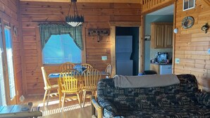 A cozy cabin interior with a wooden ceiling, a dining area with a table and chairs, a patterned sofa, and a kitchen area visible through an open doorway. - Cozy 3 Bed 2 Bath Cabin located next to Three Bears Resort in Warrens Wisconsin  (Warrens)