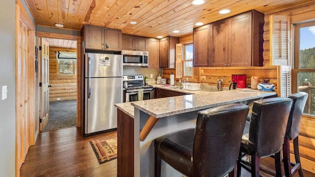 A modern kitchen with wooden cabinets, stainless steel appliances, and a bar area with leather stools. - Amazing Secluded Cabin On 10 Acres, Close To Breck (Fairplay)