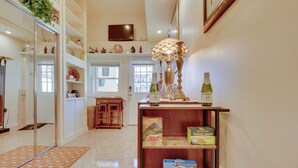 Un pasillo bien iluminado con piso de madera, una puerta de vidrio que da a una habitación y un estante con diversos objetos, incluyendo una botella, una caja de Swiss Miss chocolate con leche y una caja de té Matcha.