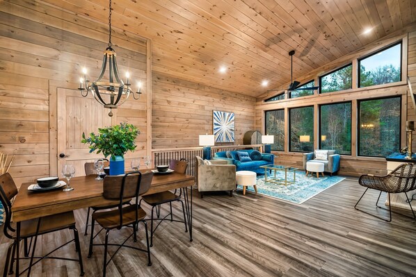 A wooden interior with a dining area, sofas, and large windows.