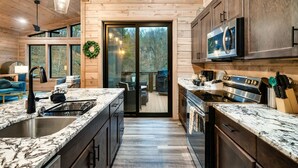 A modern kitchen with wooden cabinets, stainless steel appliances, and a large window overlooking a deck. - Perfect Modern Cabin for Couples Hot Tub Indoor Fireplace Pet friendly (Sevierville)