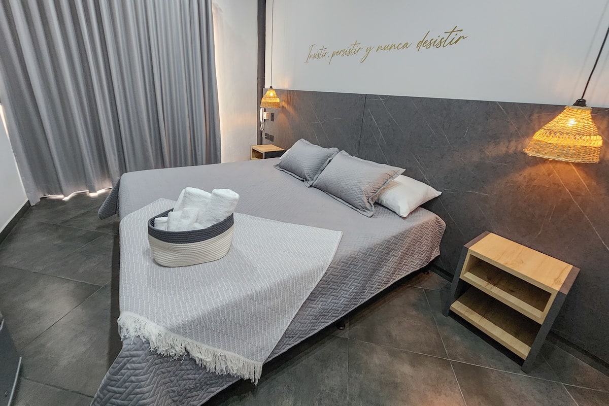 Een moderne slaapkamer met een bed, kussens, een nachtkastje, een wandlamp en een citaat op de muur.
