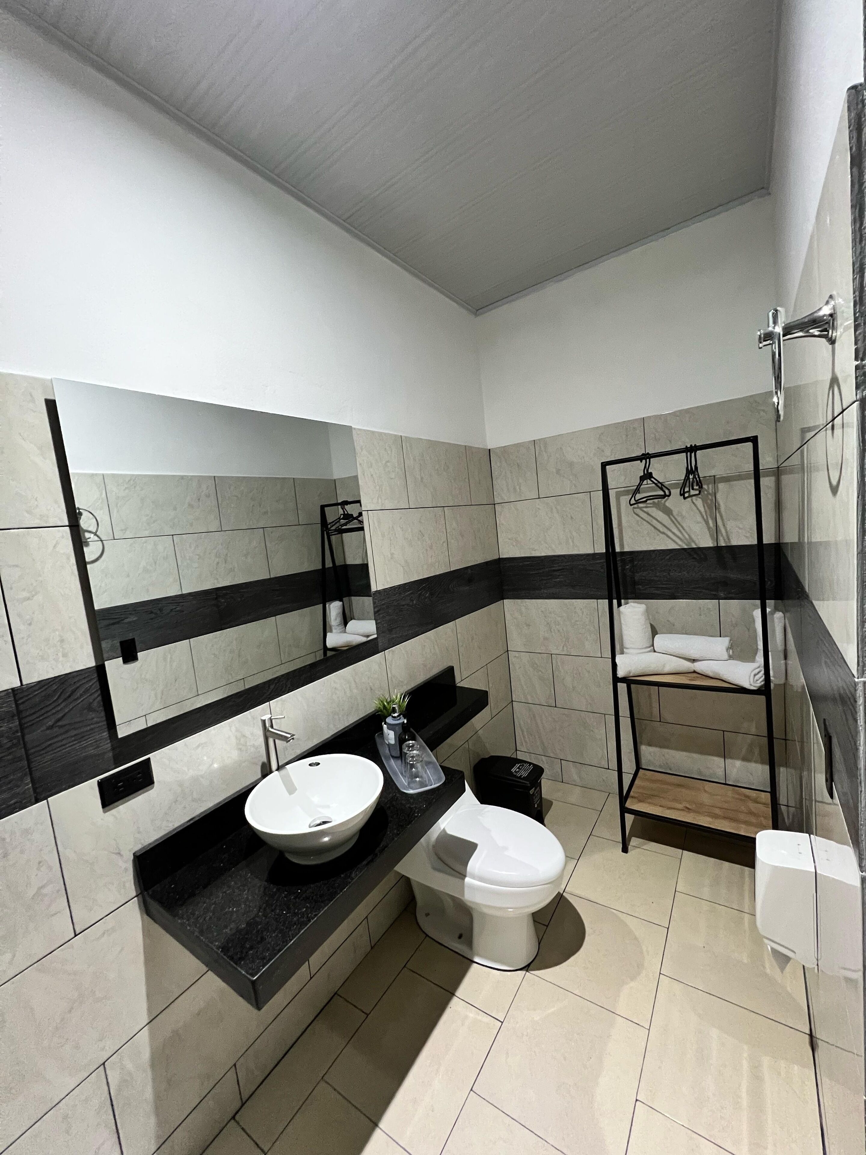Ein modernes Badezimmer mit schwarzem Waschtisch, weißem Waschbecken und Toilette. Über dem Waschbecken befindet sich ein Spiegel und darüber ein Ablagefach mit Handtüchern.
