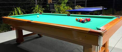 A pool table with balls and cues, surrounded by plants and a fence.