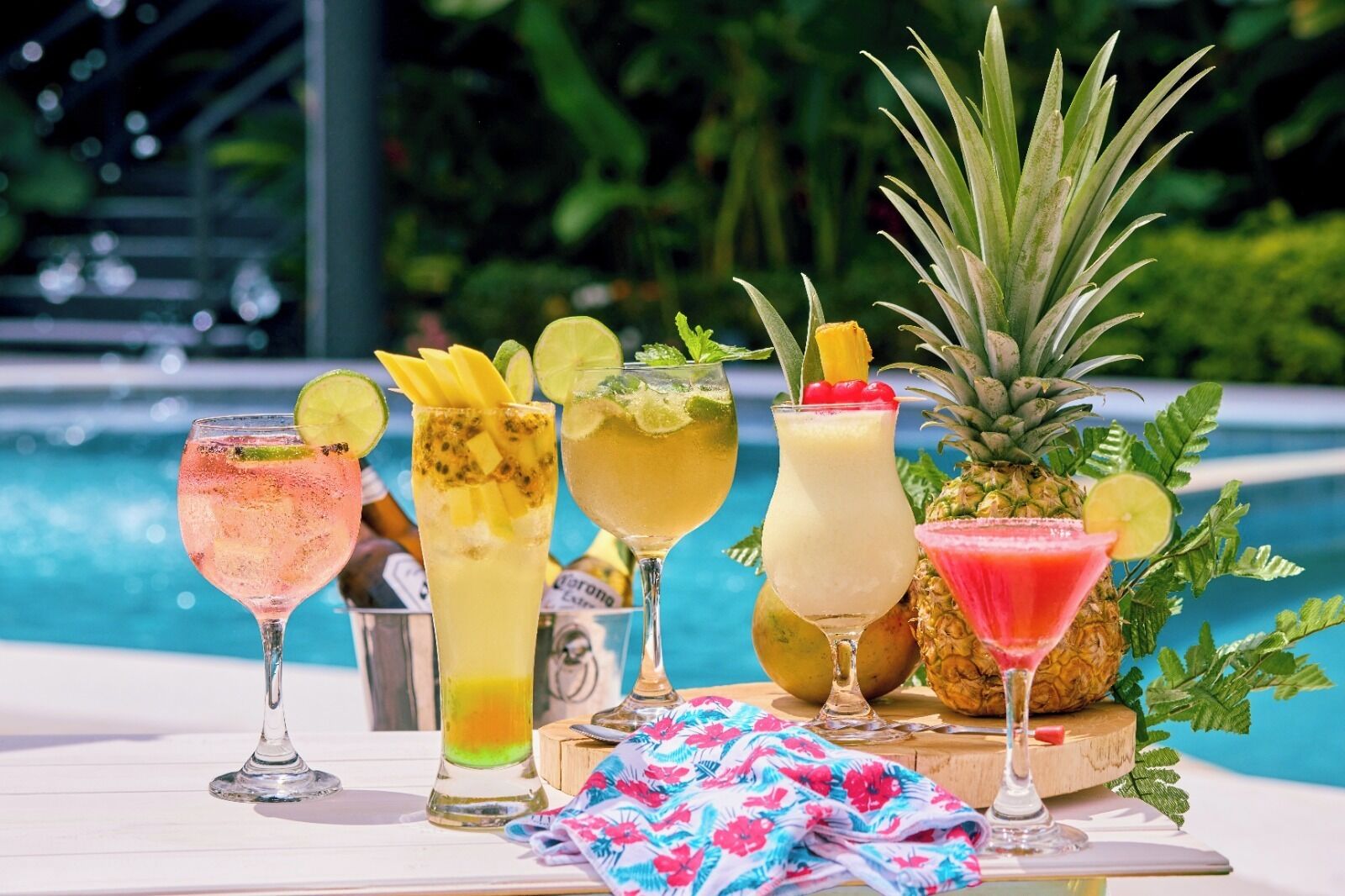 Une table où sont disposés différents cocktails tropicaux, un ananas et, au fond, une piscine.