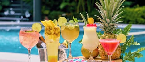 A table with various tropical cocktails, a pineapple, and a pool in the background.