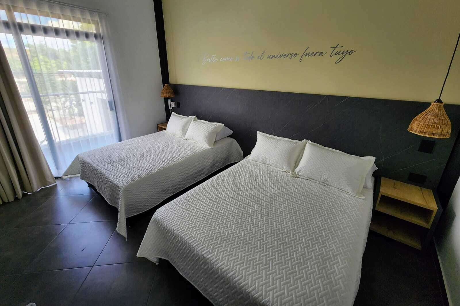A hotel room with two beds, a large window with blinds, a wall with a motivational quote, and a hanging lamp.
