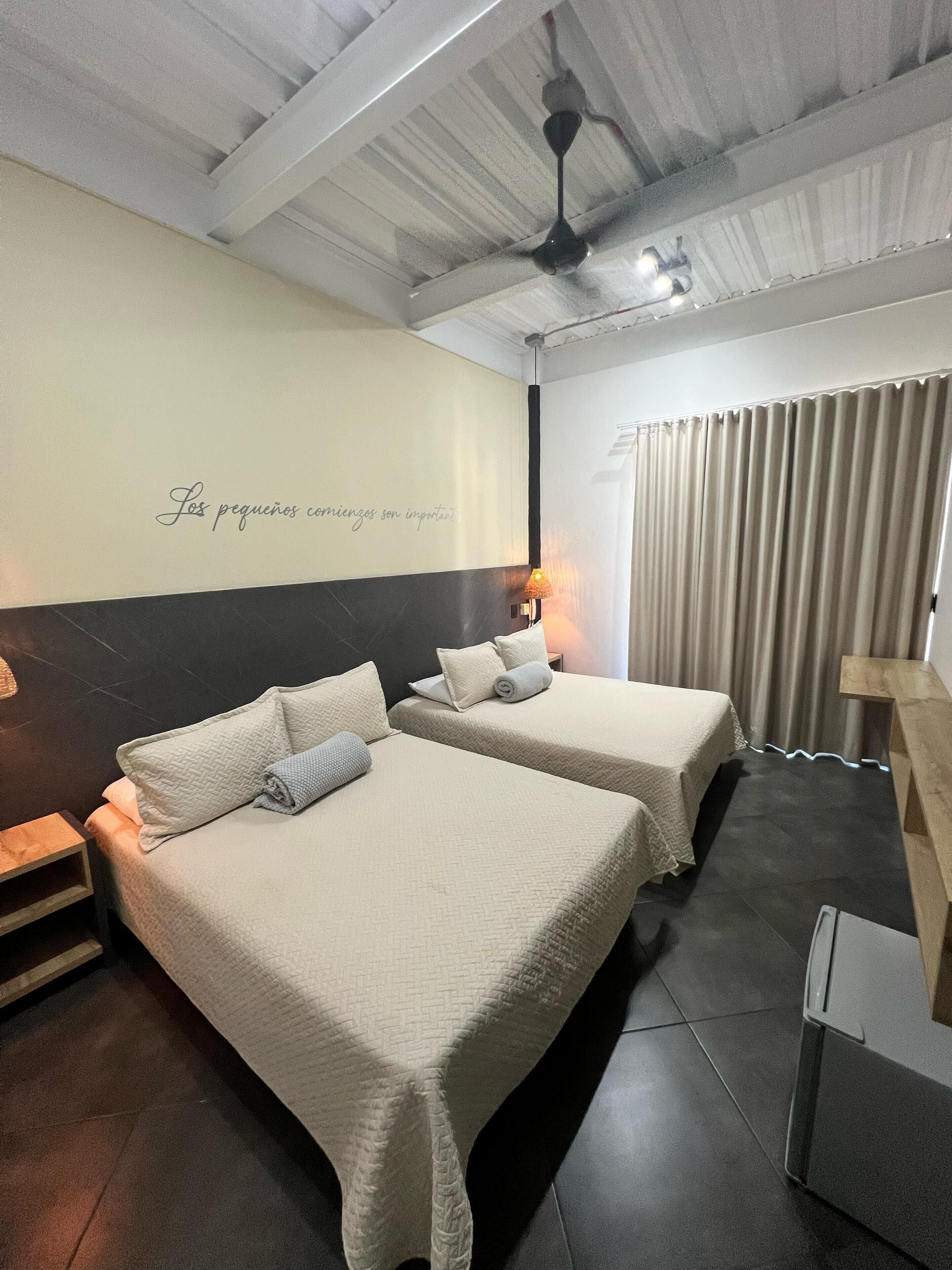 A hotel room with two beds, a ceiling fan, a wall with a motivational quote, and a window with curtains.