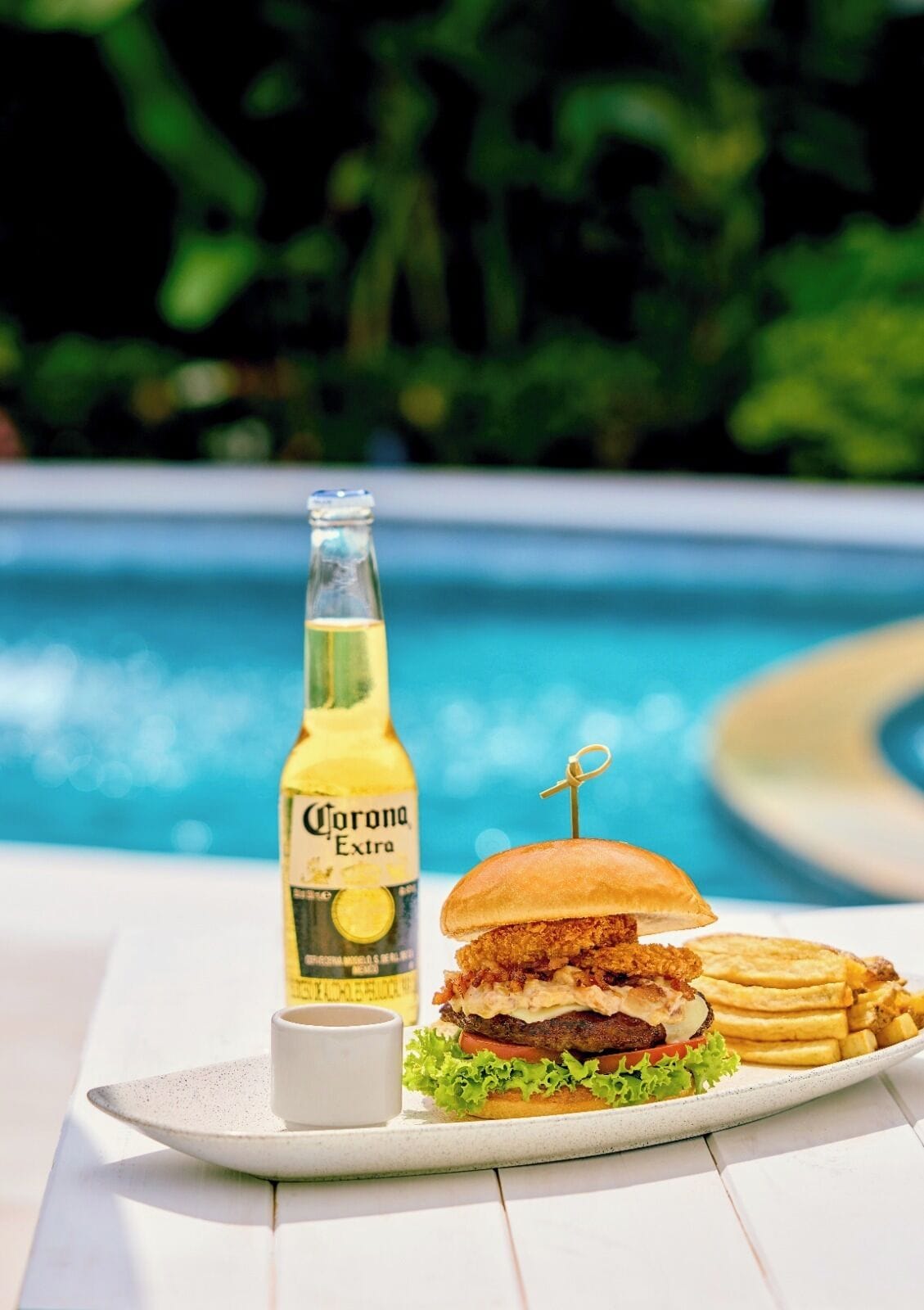 Une assiette avec un hamburger, des frites et une bouteille de bière Corona Extra, le tout placé près d’une piscine.