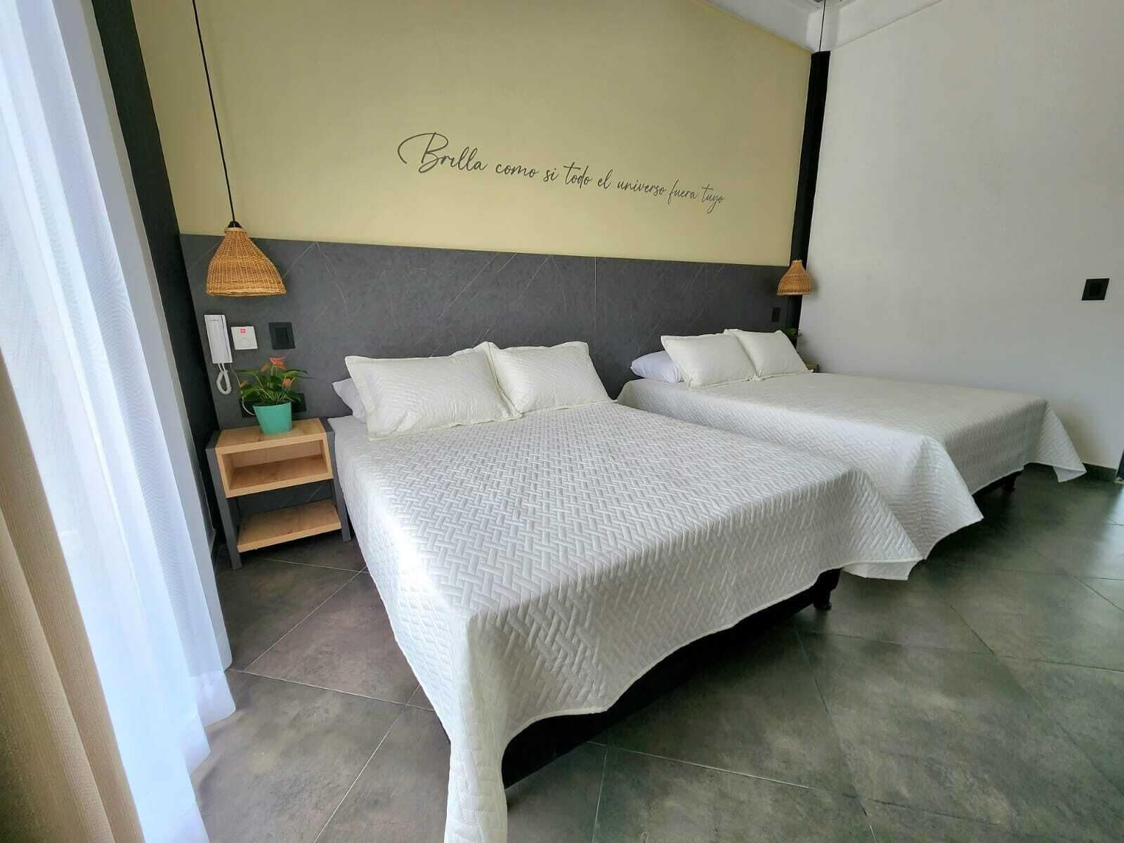 Habitación de hotel con dos camas, teléfono en la pared, una mesita con una planta y una frase decorativa en la pared.