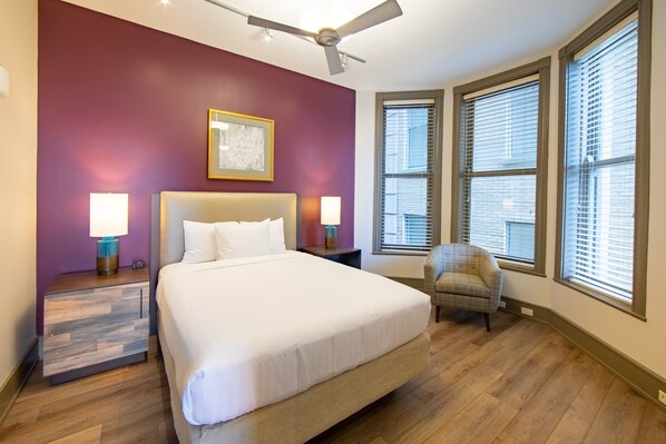 Une chambre à coucher avec un grand lit, deux tables de chevet avec des lampes, un ventilateur de plafond et une fenêtre avec des stores.
