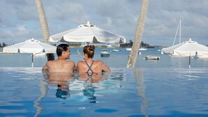 A couple in a clear blue infinity pool with white umbrellas and palm trees in the background. - Luxe 2 Bedroom Suite in Beachfront Resort (Sandys Parish)
