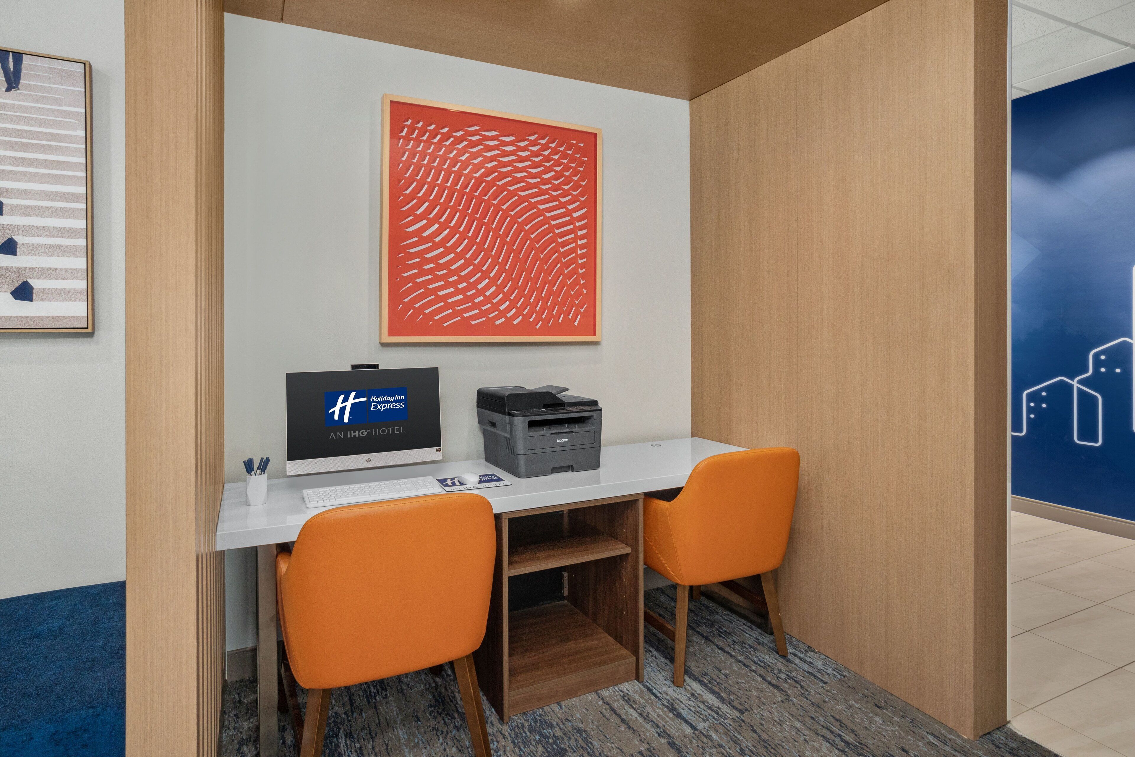 Une chambre d’hôtel équipée d’un bureau, de deux fauteuils orange, d’une imprimante et d’un grand tableau abstrait accroché au mur.