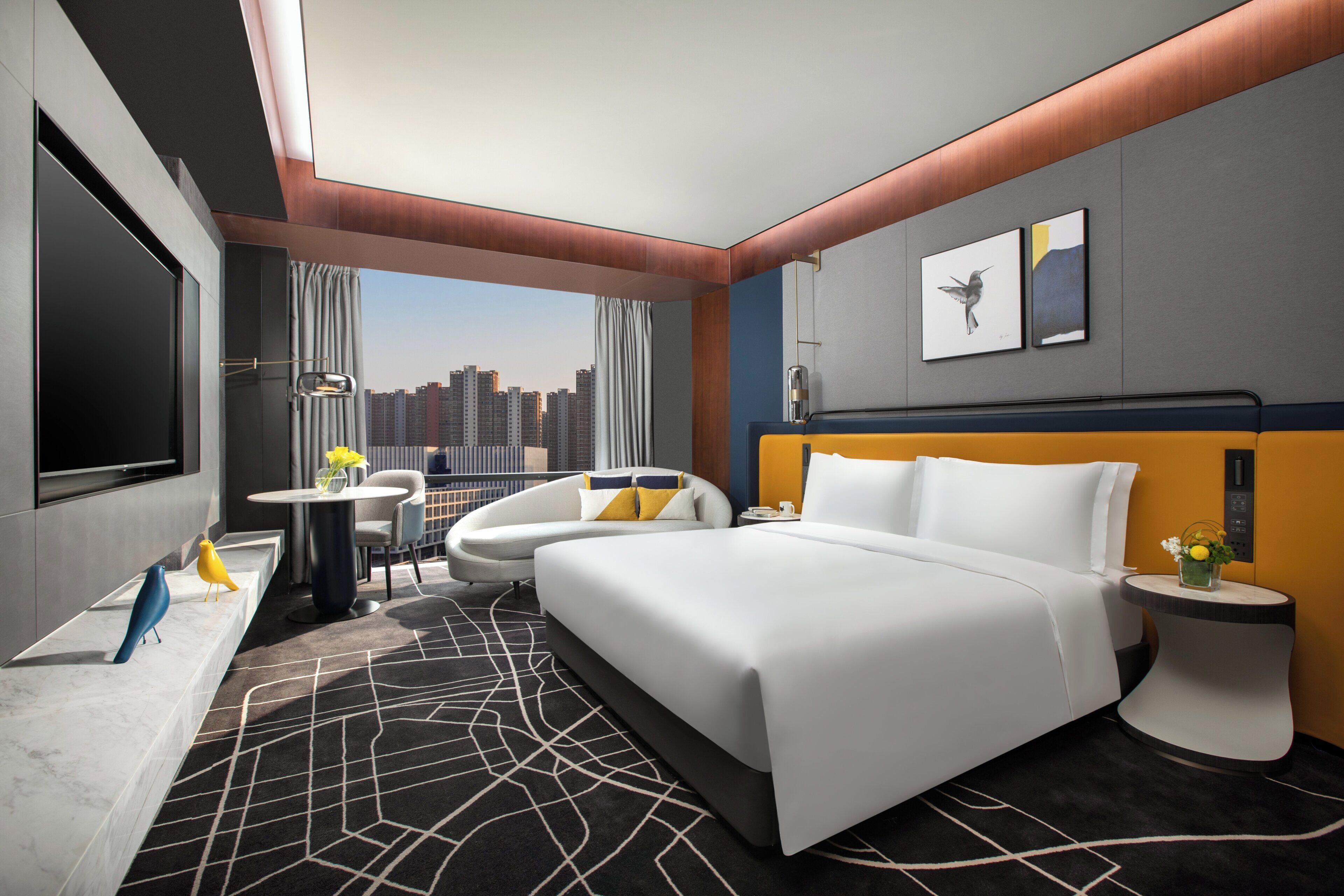 Habitació d'hotel moderna amb un llit gran, una zona de seients amb una cadira i una taula, un escriptori amb una làmpada i vista a la ciutat.