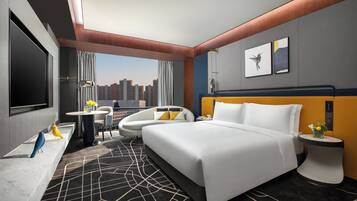 Habitació d'hotel moderna amb un llit gran, una zona de seients amb una cadira i una taula, un escriptori amb una làmpada i vista a la ciutat.