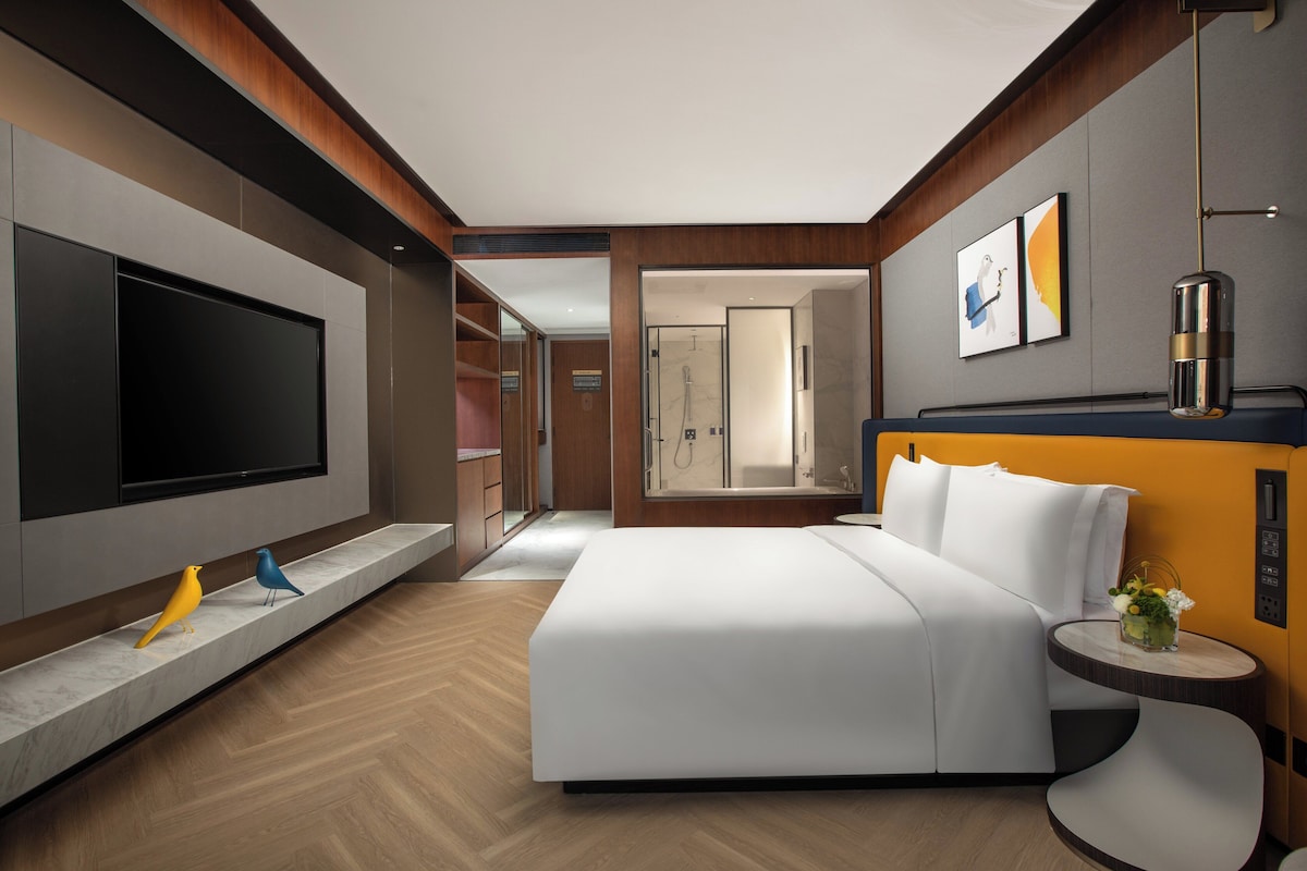 A modern hotel room with a large bed, a flat-screen TV, a small table with a vase, and a shower area.