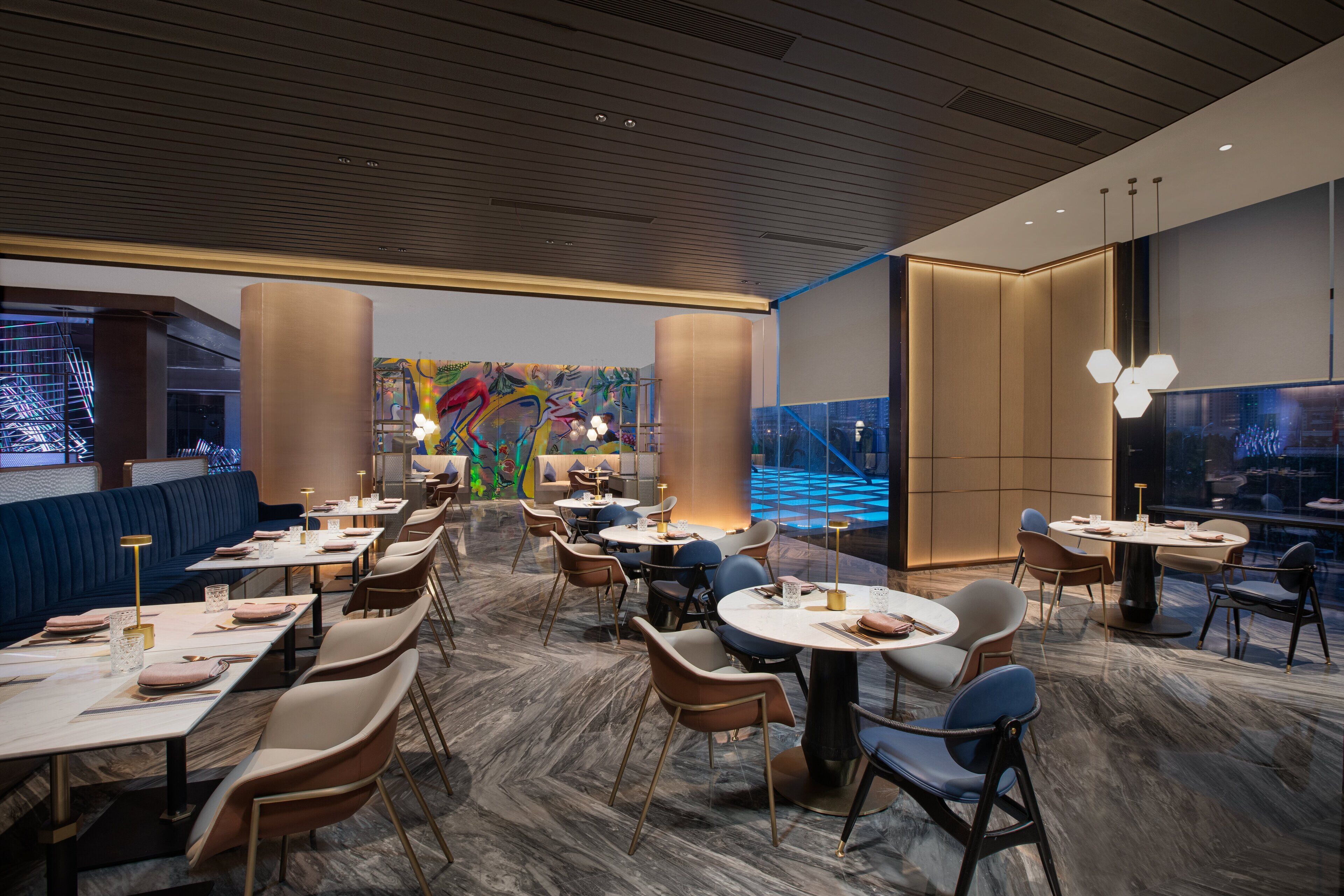 A modern restaurant with round tables and chairs, a colorful mural, and a view of the cityscape.