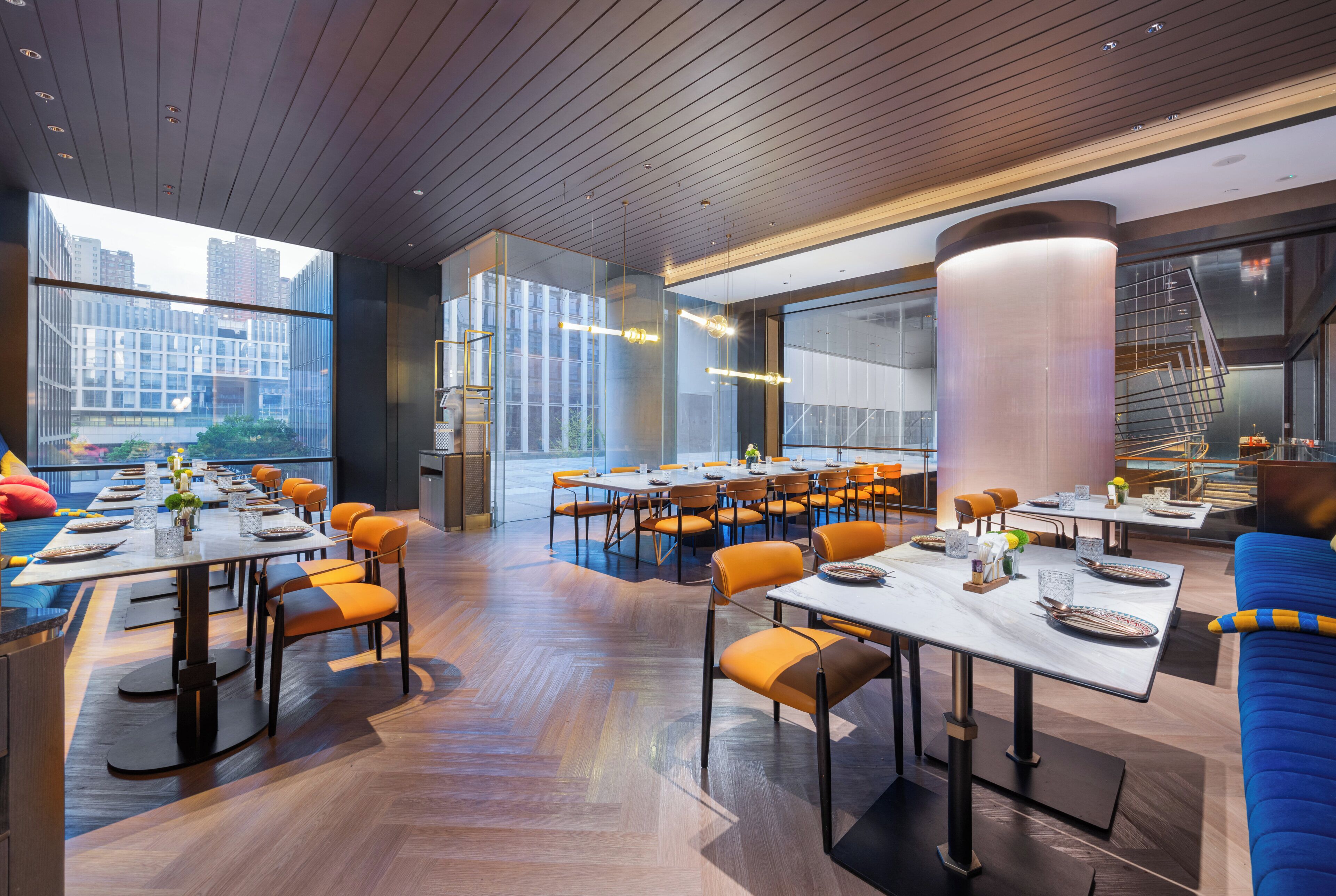 Un restaurante moderno con varias mesas de comedor y sillas anaranjadas, amplias ventanas que ofrecen vistas a la ciudad y un techo de madera.
