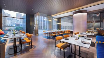 Un restaurante moderno con varias mesas de comedor y sillas anaranjadas, amplias ventanas que ofrecen vistas a la ciudad y un techo de madera.