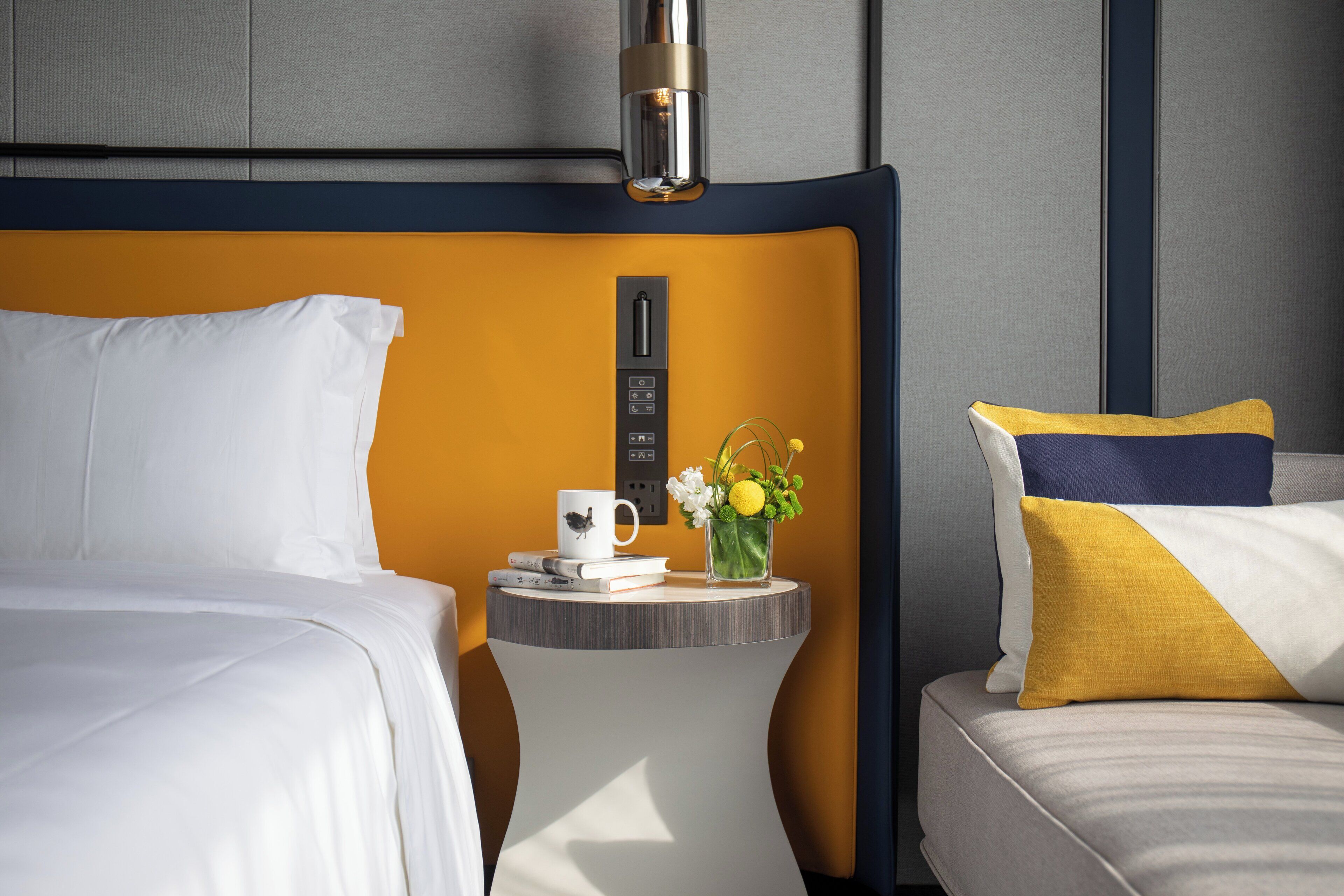 Una habitación de hotel con una cama, una mesita de noche con una taza, libros y un florero con flores.