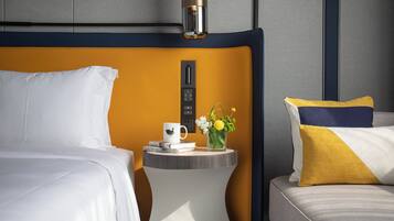 Una habitación de hotel con una cama, una mesita de noche con una taza, libros y un florero con flores.