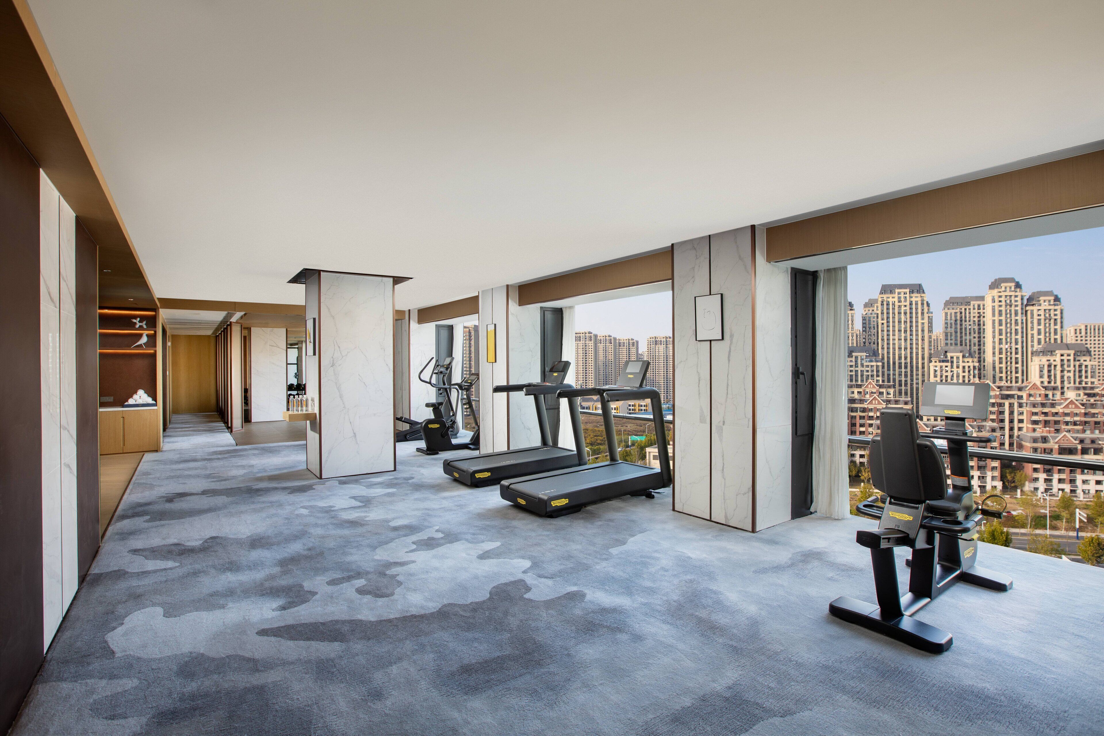 A modern hotel room with a gym area, large windows with a city view, and a minimalist design.