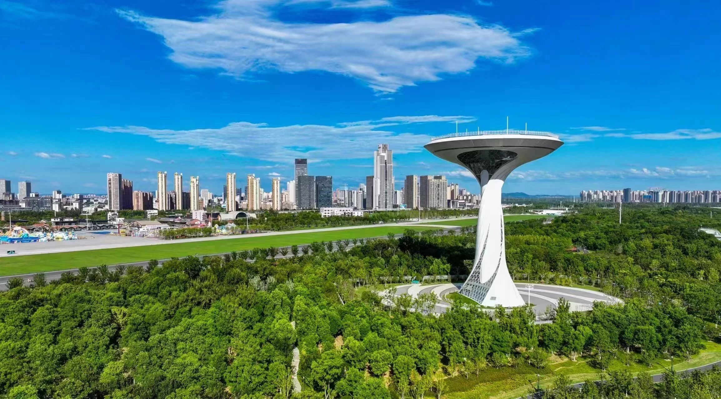 Una estructura de torre moderna en un parque urbano, con un horizonte urbano denso al fondo.