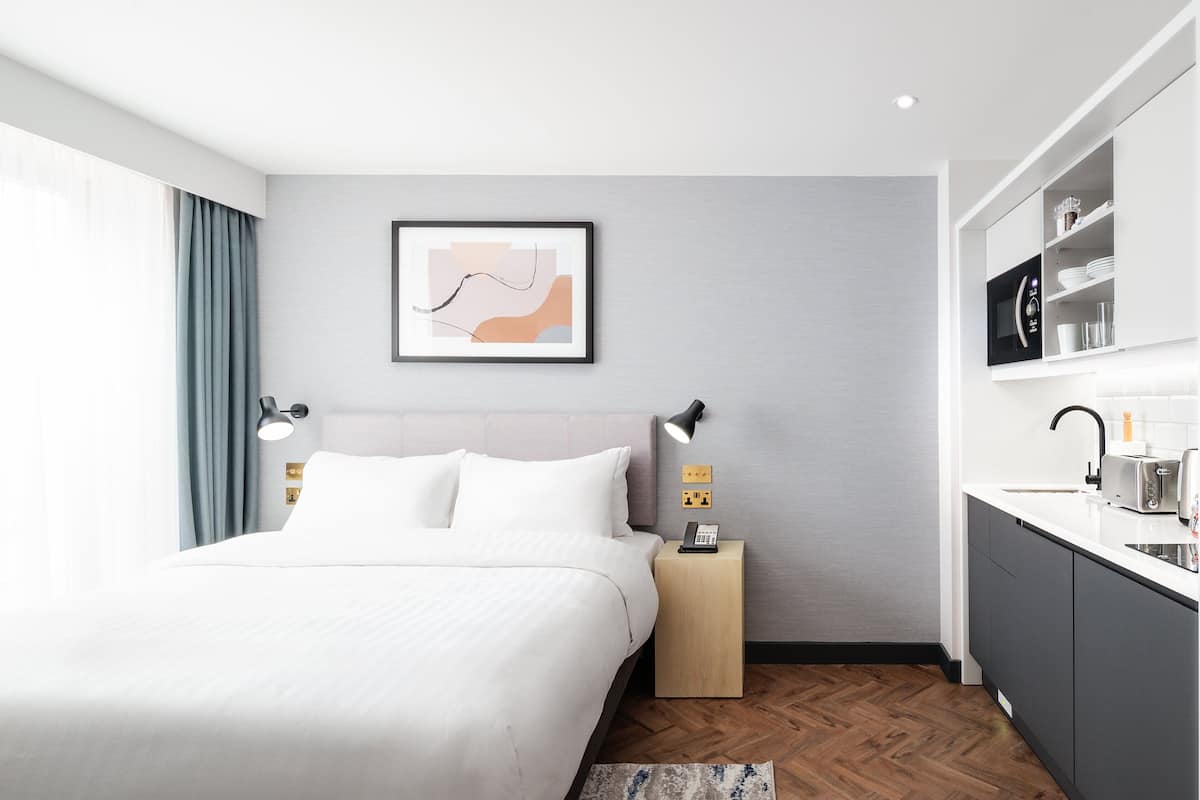 A modern hotel room with a bed, bedside table, lamp, artwork, and a kitchenette with a sink and microwave.