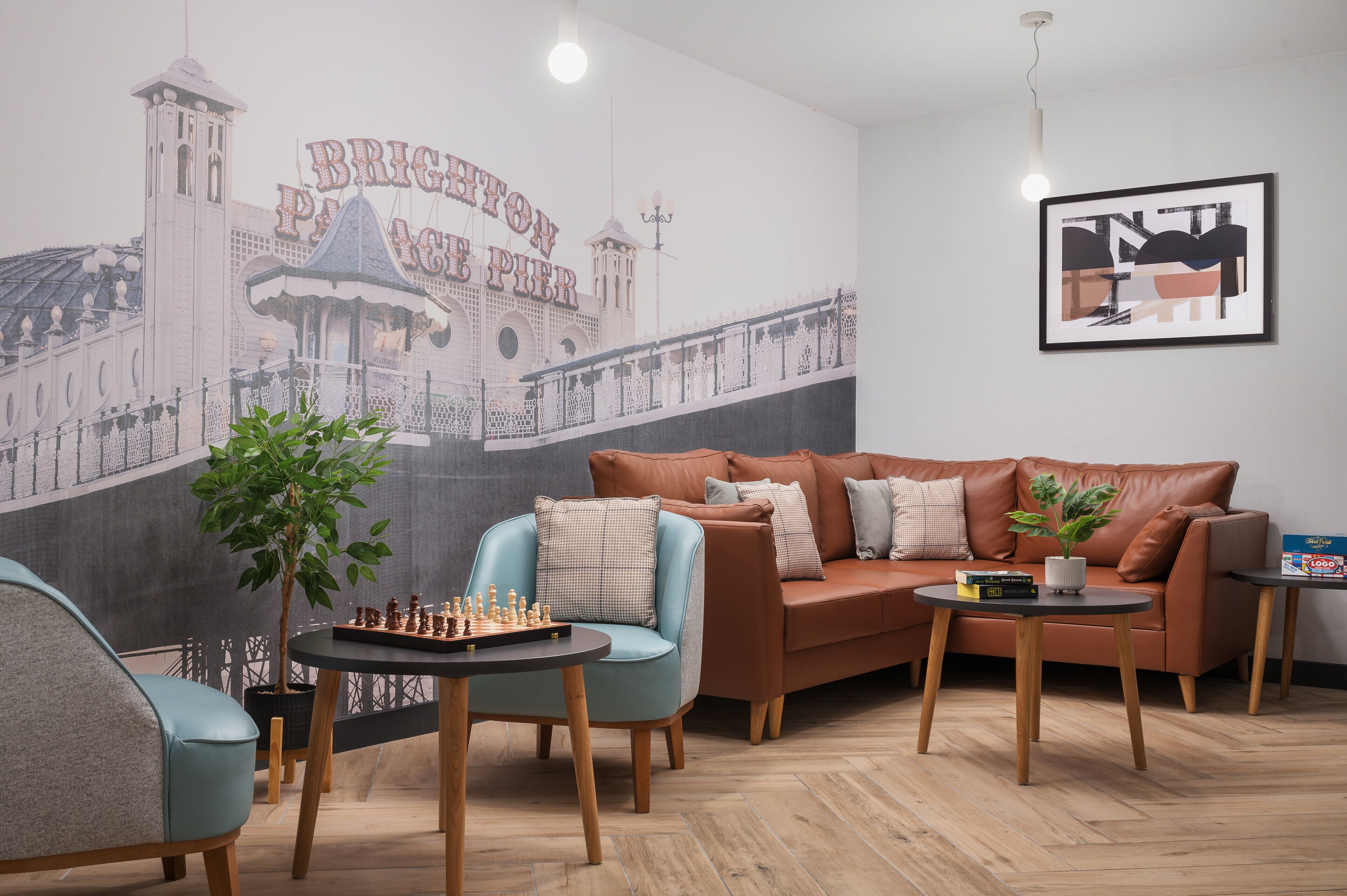 A cozy lounge area with a large mural of Brighton Pier, a brown leather sofa, a blue armchair, a chessboard, and a small table with a plant.