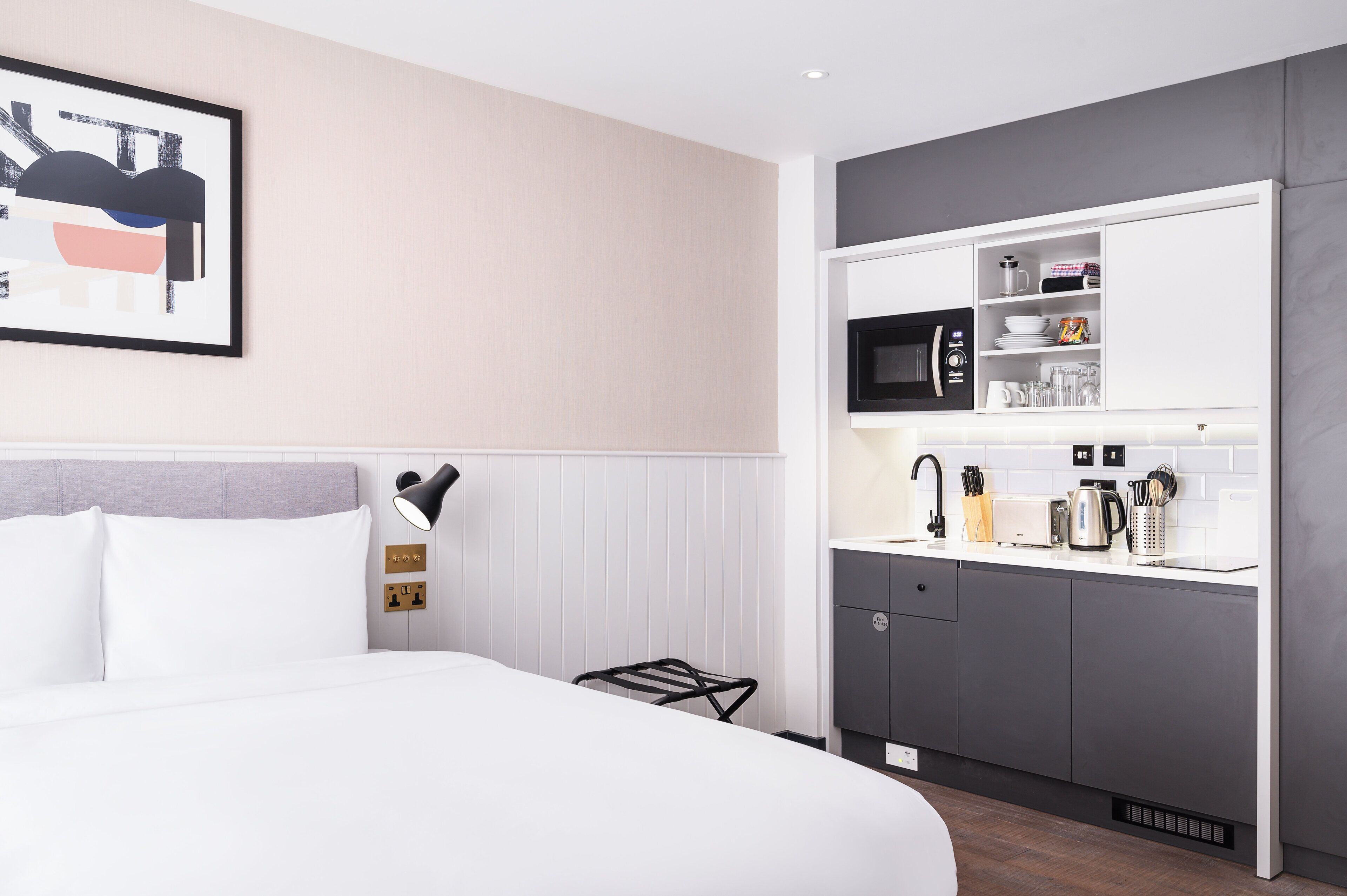 A modern hotel room with a neatly made bed, a wall-mounted artwork, a kitchen area with appliances, and a towel rack.