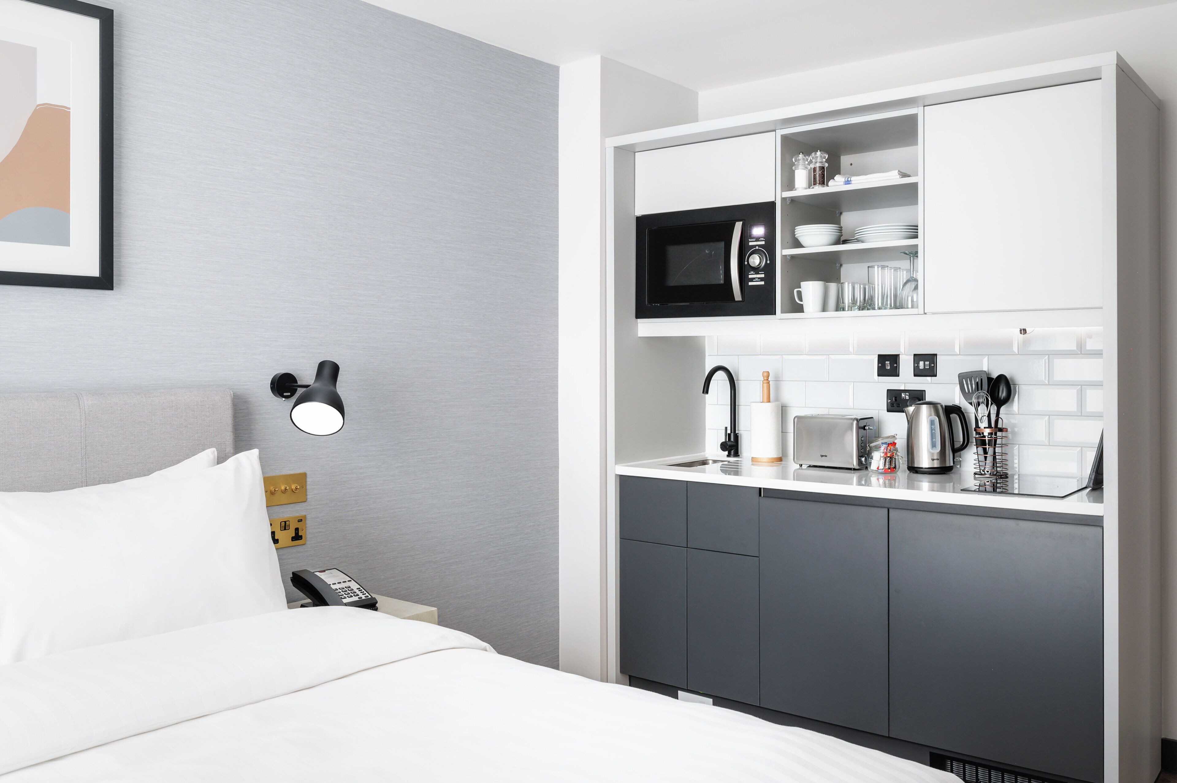 A hotel room with a bed, a bedside table with a lamp, a microwave, a toaster, and a coffee maker on the countertop.