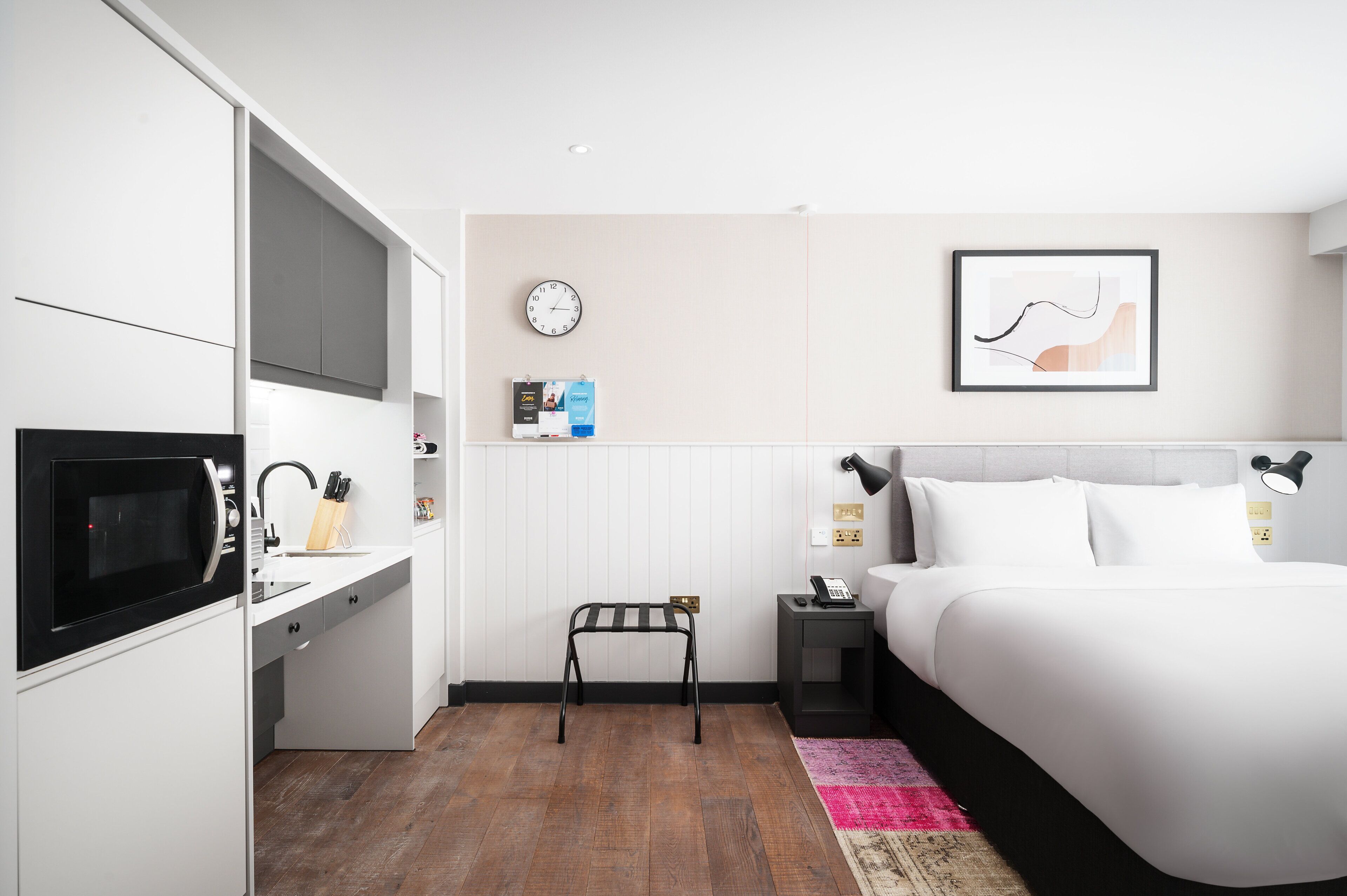 Ett modernt hotellrum med ett litet kök, en säng, ett sängbord, en klocka och en inramad tavla på väggen.