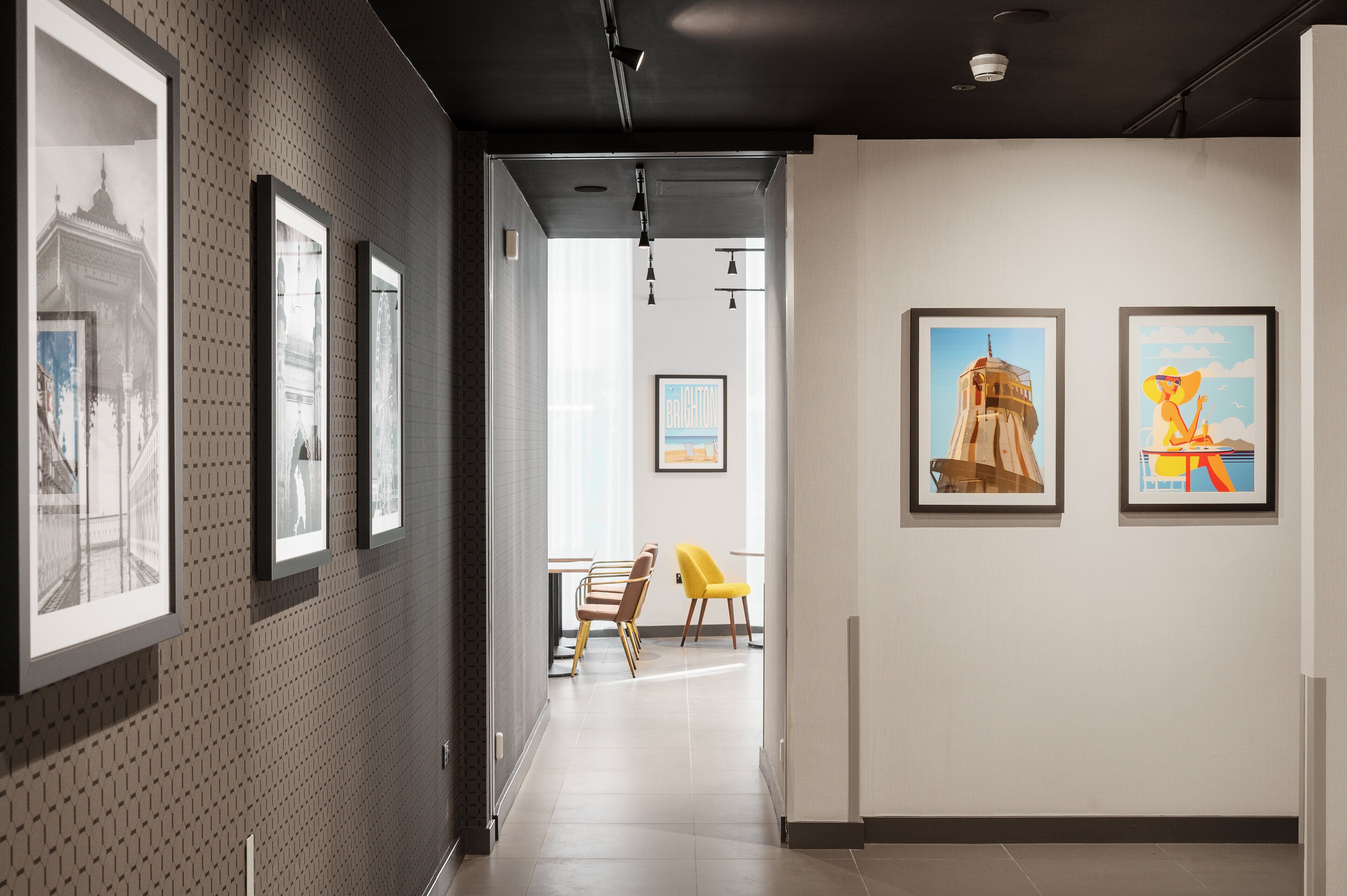 Un pasillo con dibujos arquitectónicos enmarcados a la izquierda, una zona de comedor con sillas y una mesa en el centro, y dos carteles coloridos en la pared derecha.